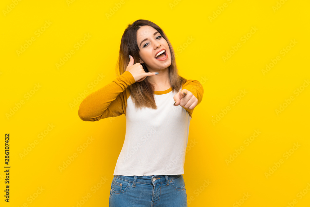 Pretty young woman over isolated yellow wall making phone gesture and pointing front Pretty young woman over isolated yellow wall making phone gesture and pointing front