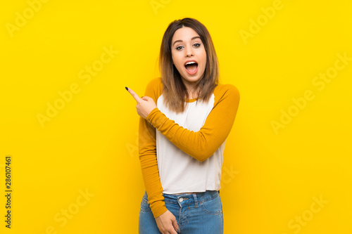 Pretty young woman over isolated yellow wall surprised and pointing side