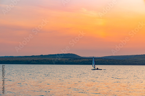 atmospheric European lake scenery landscape in vivid orange sunset evening time with small sail boat on slightly wavy water surface and forest mail land background 