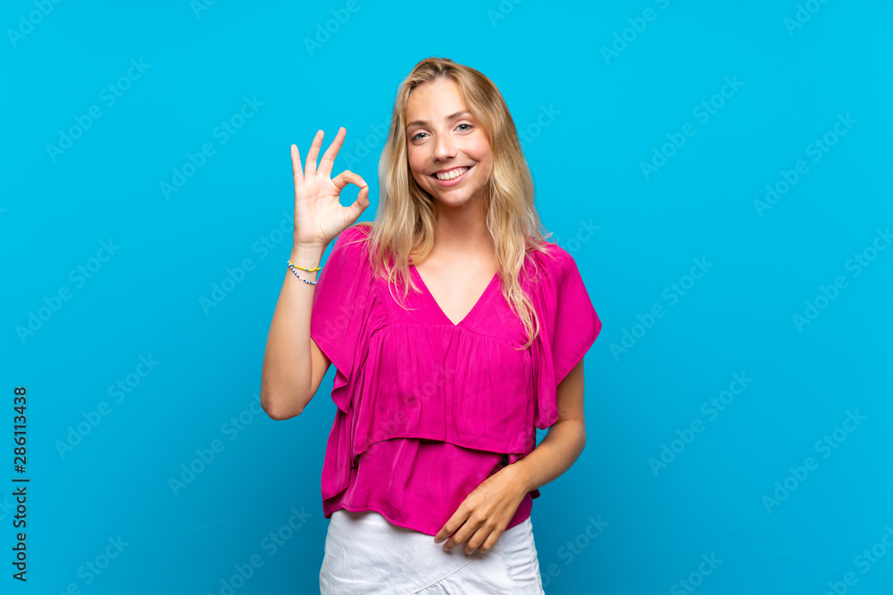 Blonde young woman over isolated blue background showing ok sign with fingers Blonde young woman over isolated blue background showing ok sign with fingers