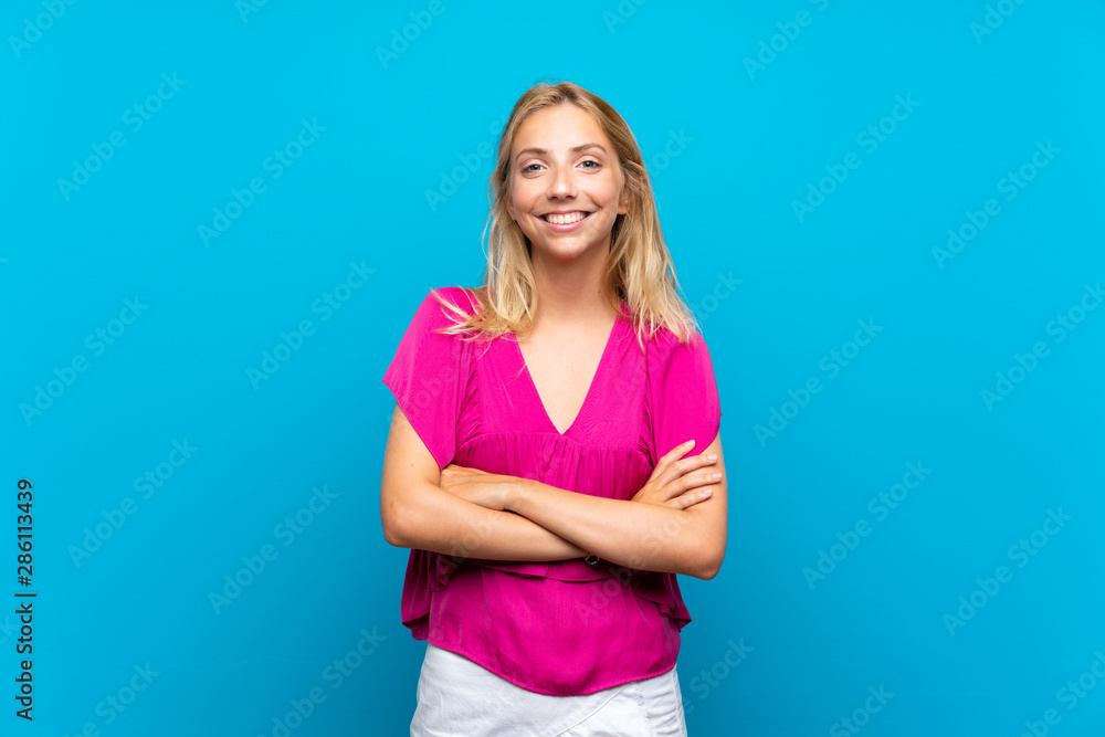 Blonde young woman over isolated blue background keeping the arms crossed in frontal position Blonde young woman over isolated blue background keeping the arms crossed in frontal position