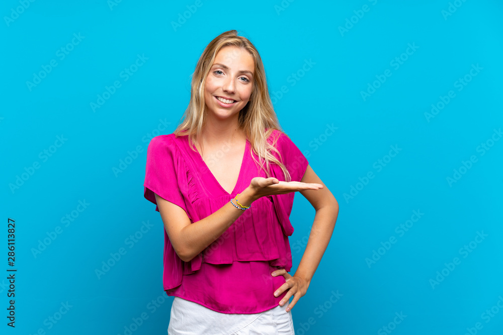 Blonde young woman over isolated blue background presenting an idea while looking smiling towards Blonde young woman over isolated blue background presenting an idea while looking smiling towards