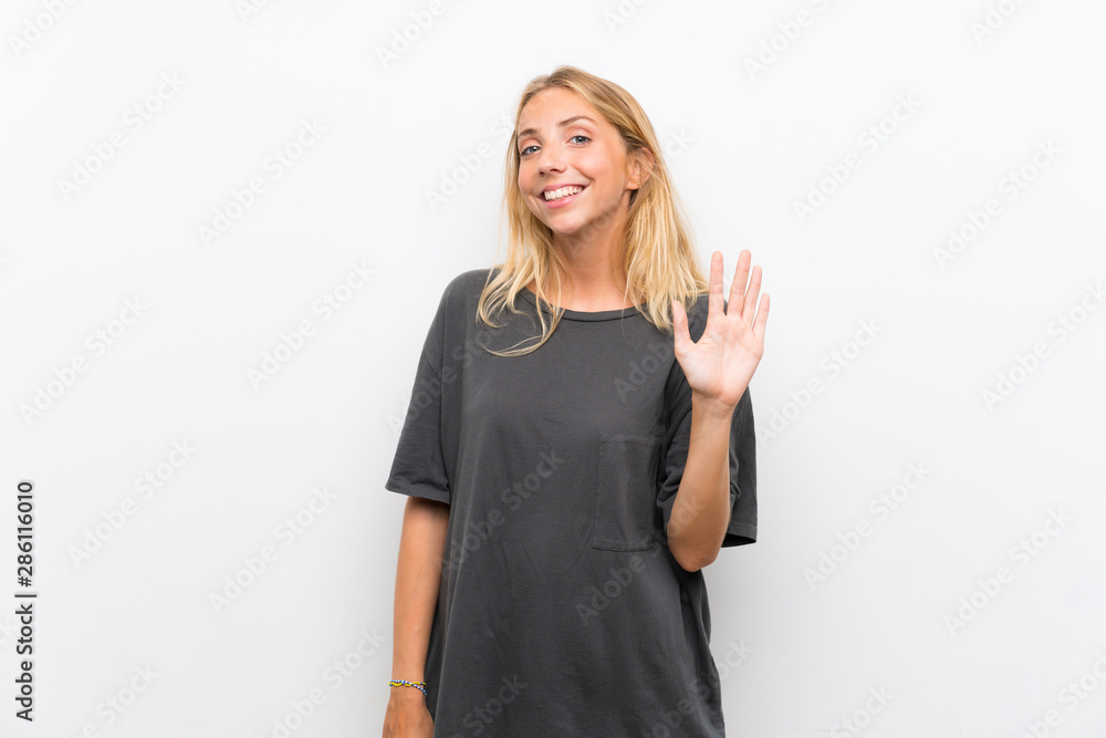 Blonde young woman over isolated white background saluting with hand with happy expression Blonde young woman over isolated white background saluting with hand with happy expression
