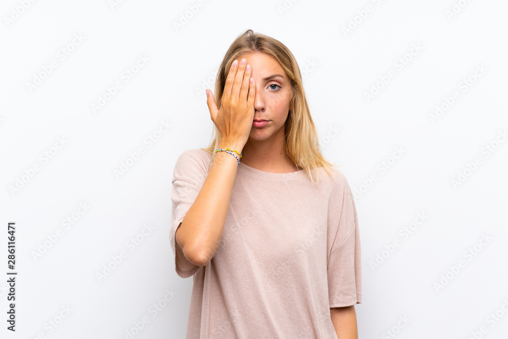 Blonde young woman over isolated white background covering a eye by hand Blonde young woman over isolated white background covering a eye by hand