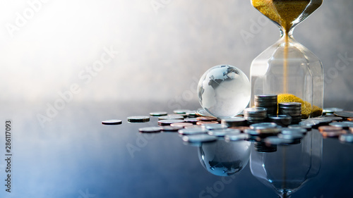 Global business investment growth. Wealth concept. Gold sand running through the shape of modern hourglass with world globe crystal glass and US dollar coins on blue reflective table. photo