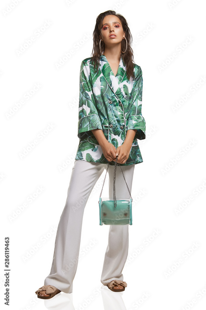 Full shot of a young chestnut European lady dressed in white casual trousers, loose jacket with white and green jungle print and beige sandals with a wide textile bow. Full shot of a young chestnut European lady dressed in white casual trousers, loose jacket with white and green jungle print and beige sandals with a wide textile bow.