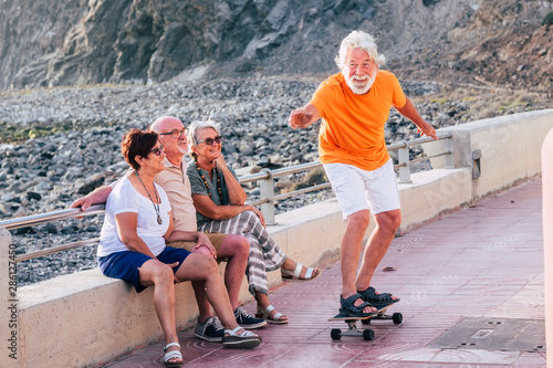 group of seniors and mature people at the beach have fun looking at old man riding a skateboard and laughing with scare face - woman touching the man photo