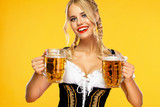 Young sexy oktoberfest girl waitress, wearing a traditional Bavarian or german dirndl, serving two big beer mugs with drink isolated on yellow background.
