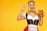 Young sexy Oktoberfest girl waitress, wearing a traditional Bavarian or german dirndl, serving big beer mugs with drink isolated on yellow background. Woman showing ok sign with fingers.