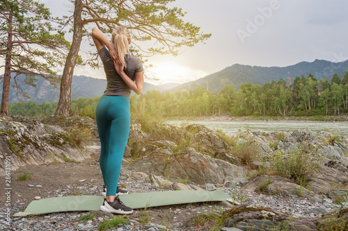 Blond pretty woman with sexy tight booty is doing stretching by stretching joining hands behind your back against the mat in the picturesque place in mountains near the river and tree on stones.