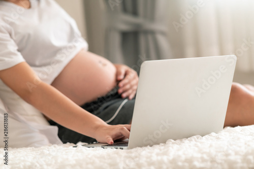 Asian pregnant woman using laptop in bed. Happy smile. With copy space. Side shot.