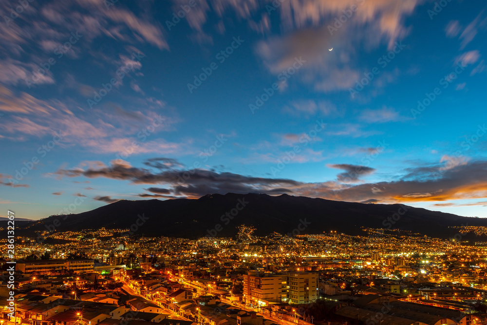 Long exposure cityscape of Quito at night with a crescent moon and the impressive active Pichincha volcano in the background, Ecuador.