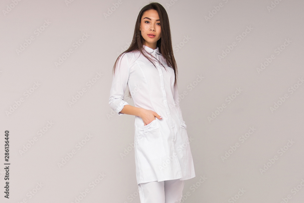 young stylish happy nurse with long brunette hair is standing with hand in her pocket, looking straight and smiling on white wall background. medical fashion concept. free space young stylish happy nurse with long brunette hair is standing with hand in her pocket, looking straight and smiling on white wall background. medical fashion concept. free space