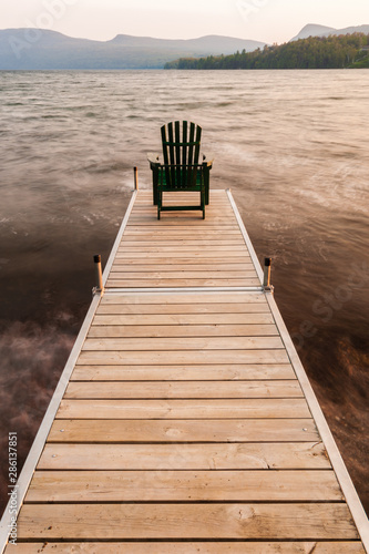 Photo Adirondack chair on the end of a dock on a lake.