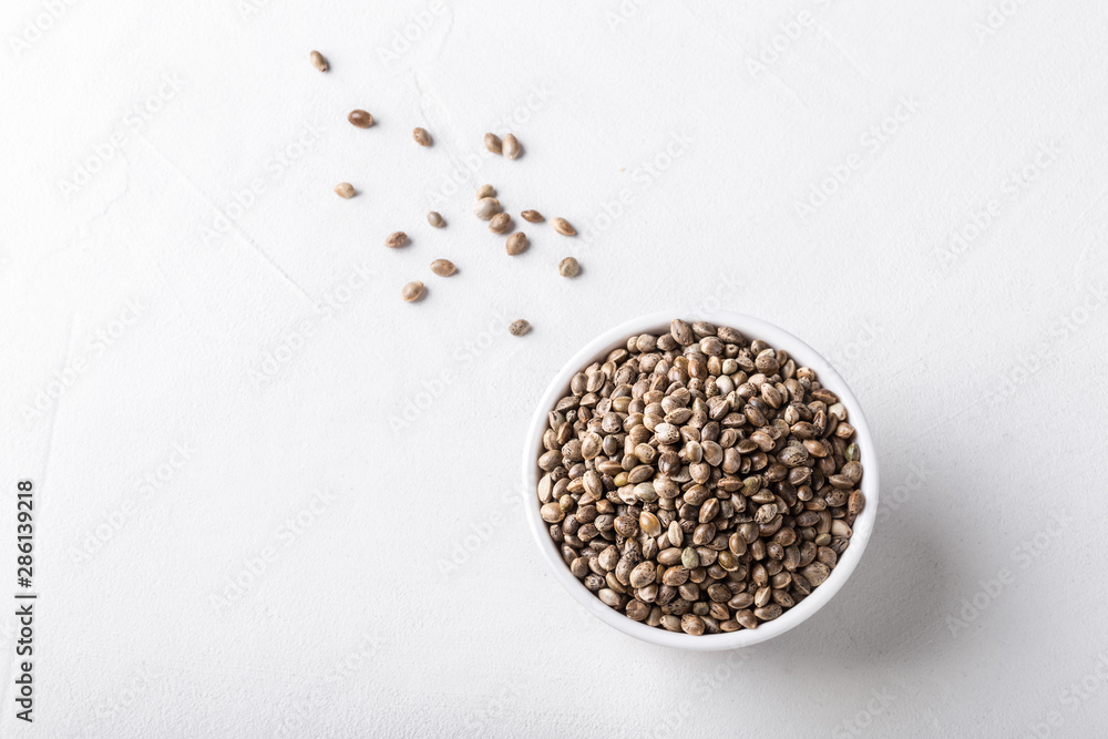 Raw organic unrefined hemp seeds in small white bowl on white concrete background. Raw organic unrefined hemp seeds in small white bowl on white concrete background.