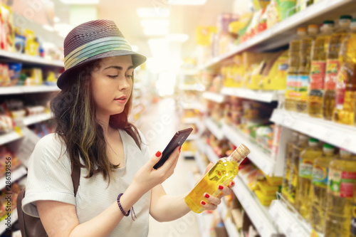 A young beautiful woman holds a bottle of oil in her hand and takes a picture of her on a mobile phone. In the background shelves with products. The concept of modern shopping in the store. Light photo