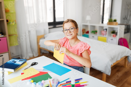 childhood, creativity and hobby concept - creative girl in glasses with color paper sitting at table at home