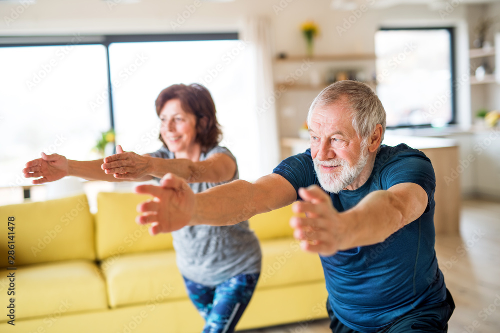 A senior couple indoors at home, doing exercise indoors. A senior couple indoors at home, doing exercise indoors.