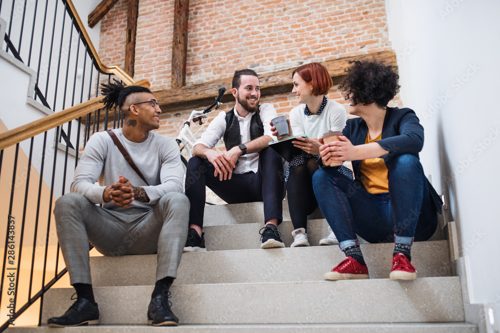 Fototapeta premium Group of young businesspeople sitting on stairs indoors, talking.