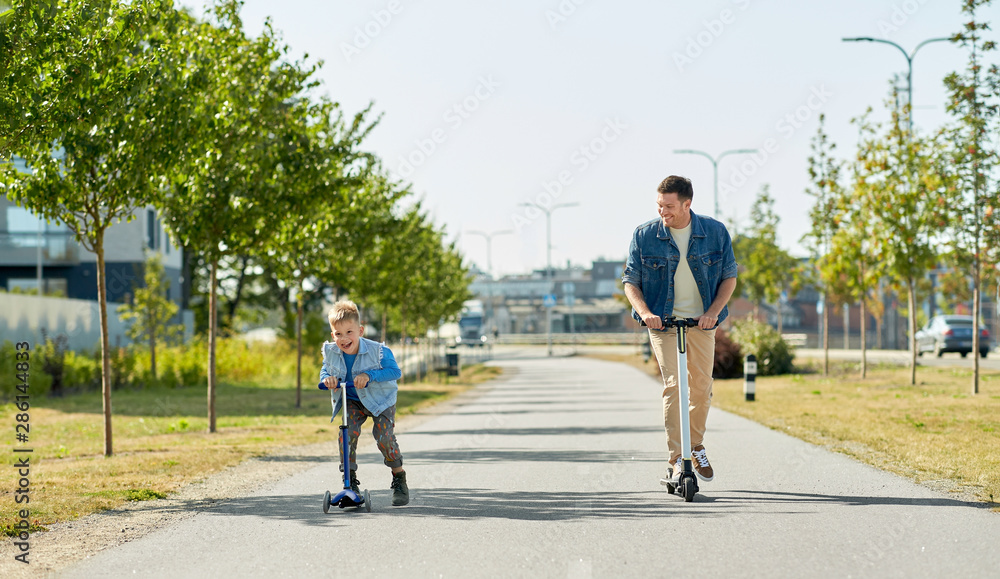 family, leisure and fatherhood concept - happy father spending time with little son riding scooters in city