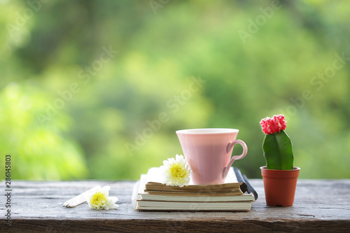 Pink cup with small cactus plant in brown pot and flowers with diary notebooks on wooden table with green nature background