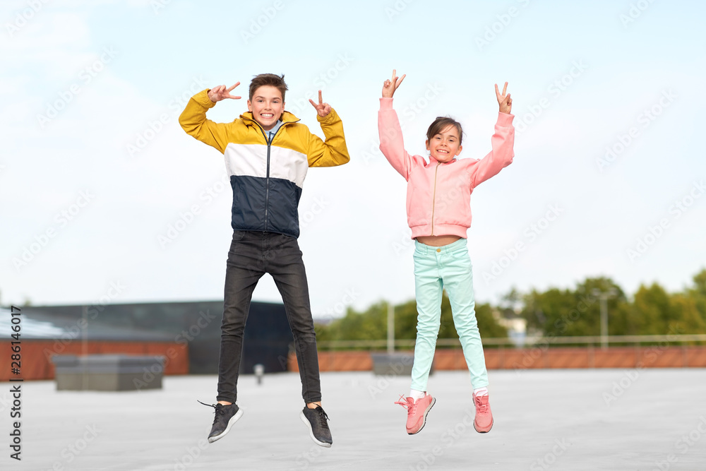 Obraz premium childhood, happiness and fun concept - happy children or brother and sister jumping on rooftop and showing peace gesture