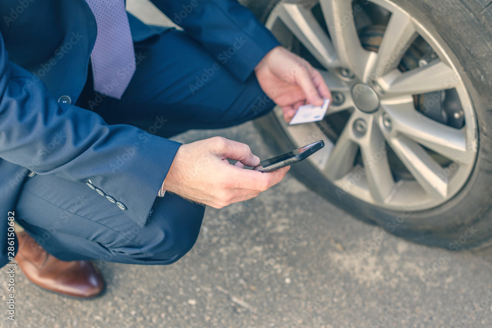 A businessman in suit crouching near his car with  punctured wheel. Man holding mobile phone and business card with data of isurance company. Concept - obrazy, fototapety, plakaty 