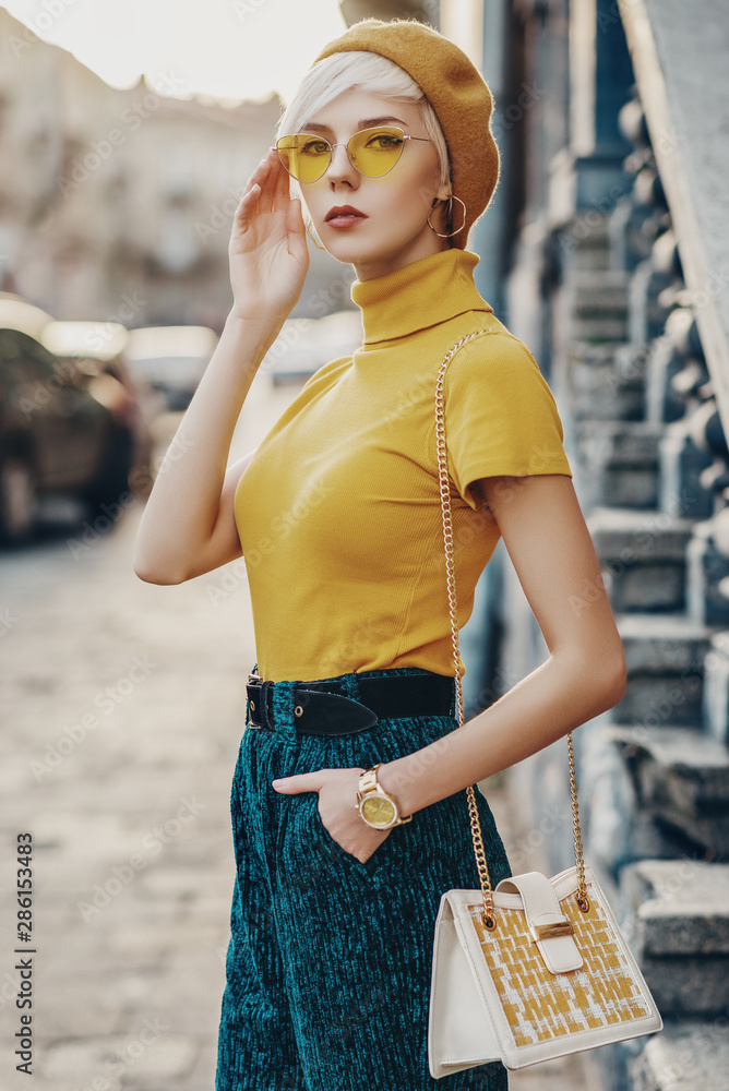Outdoor fashion portrait of young elegant lady wearing colorful outfit yellow beret glasses turtleneck green trousers golden wrist watch holding stylish white bag posing in street Stock Adobe Stock