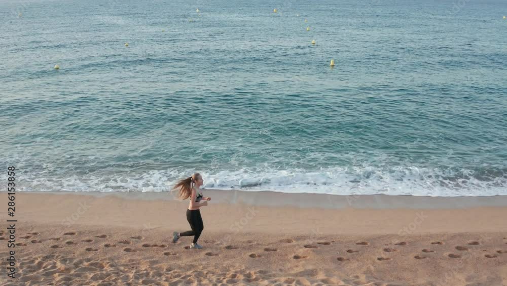 Aerial shot of long haired slim female running along sandy beach, blue waves breaking on sea shore. Side view of jogger in sporty outfit doing sports on coast against background of scenic seascape Aerial shot of long haired slim female running along sandy beach, blue waves breaking on sea shore. Side view of jogger in sporty outfit doing sports on coast against background of scenic seascape