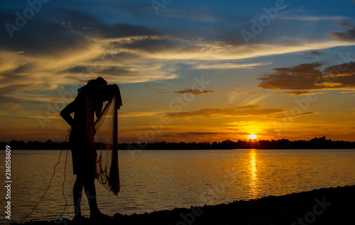 Silhouette of Thai Fisherman catching the fish in twilight, Beautiful Sunset in the sky with sky blue and orange light of the sun through the clouds in the sky, Orange and red dramatic colors.