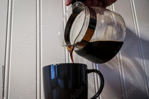 Hot fresh coffee is being poured from a glass coffee pot into a black coffee mug