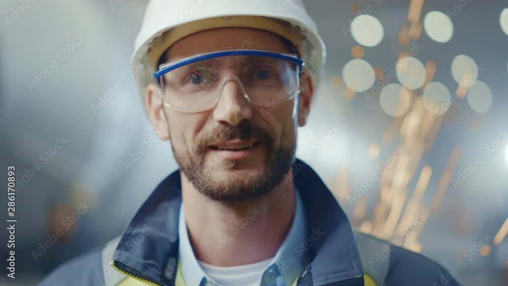 Portrait of Smiling Professional Heavy Industry Engineer / Worker Wearing Safety Uniform, Goggles and Hard Hat. In the Background Unfocused Large Industrial Factory where Welding Sparks Flying Portrait of Smiling Professional Heavy Industry Engineer / Worker Wearing Safety Uniform, Goggles and Hard Hat. In the Background Unfocused Large Industrial Factory where Welding Sparks Flying