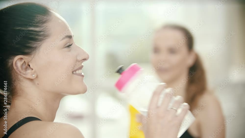 Two smiling women standing in the gym and drink water from the bottles Two smiling women standing in the gym and drink water from the bottles