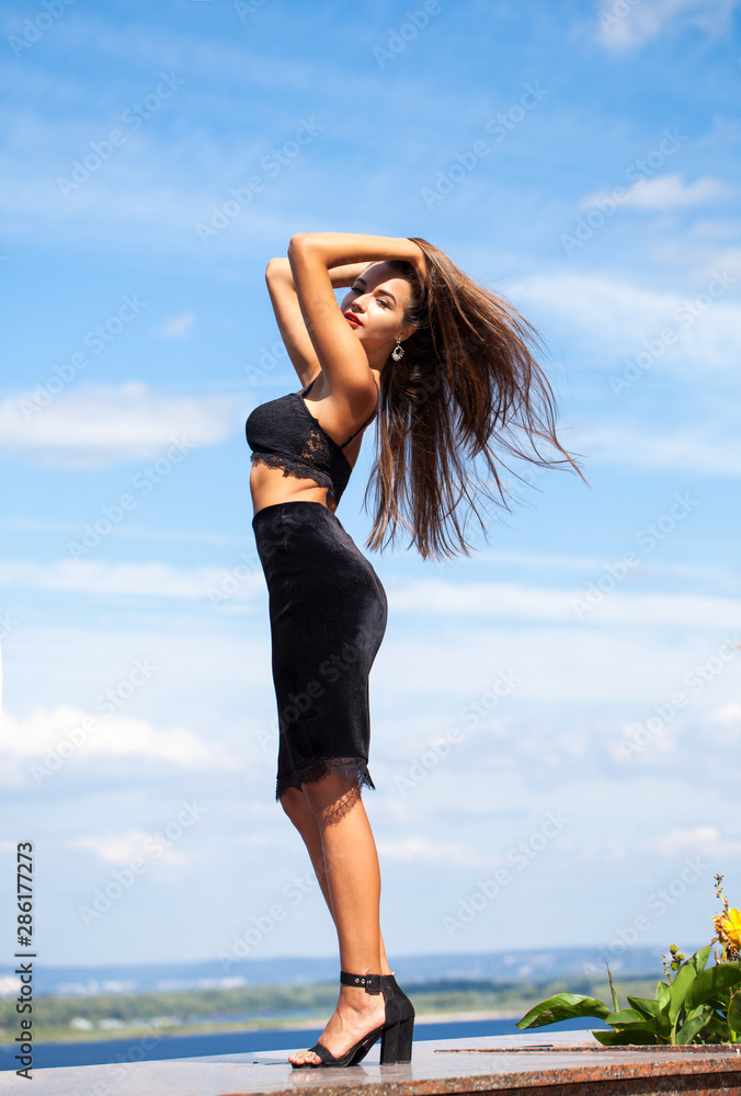 Portrait of a young beautiful girl in a black skirt and a bra posing on a background of blue sky