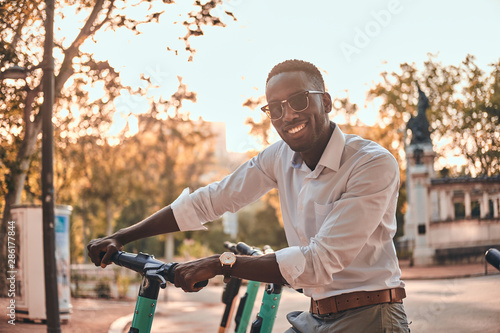 Cheerful smiling man is choosing right scooter from parking at bright sunny day.