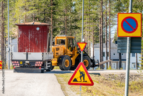 Big yellow tractor turn right while relocates old small red woden house in the trailer, sunny summer day, nearby to Umea city, North Sweden.Road construction sign at the road side
