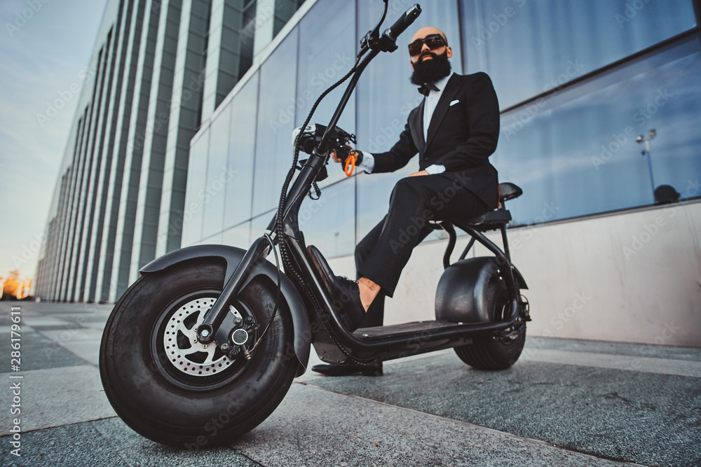 Smart attractive businessman is sitting on his electro scooter while posing for photographer.