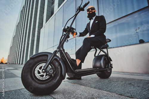 Smart attractive businessman is sitting on his electro scooter while posing for photographer.