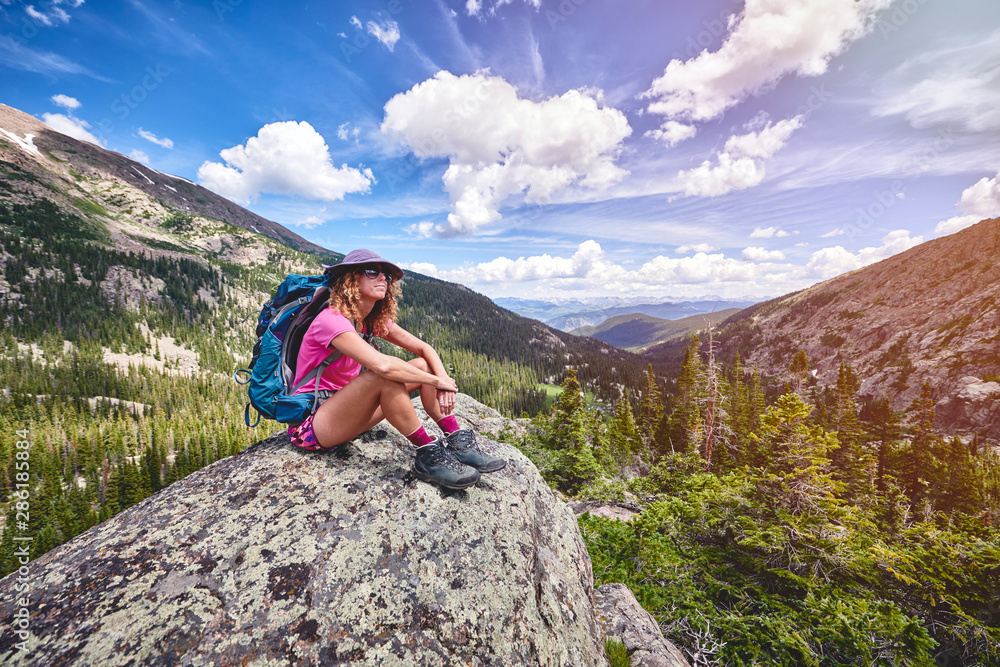 Naklejka premium A young woman carrying a backpack, sitting on top of a mountain.