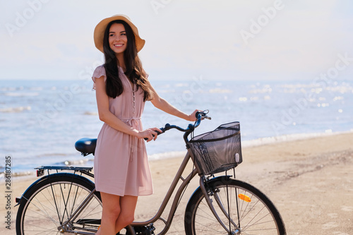 Pretty cheerful girl in hat is walking on the seaside with her bike on bright sunny day.