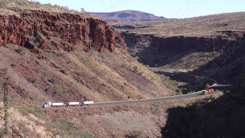 Slow moving Road train trucks driving through a valley on outback highway Western Australia, with mountain range in the background.