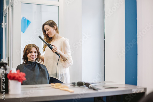 beauty, hairstyle concept, happy young woman and hairdresser with hair iron making hairdo at hair salon. Woman Having Hair Stylized Hairdresser. Smoothes Hair Curling. Stylist Using Tool for Modeling