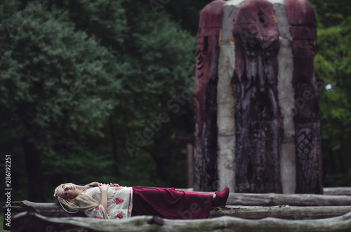 Mysterious young woman in folk costume lies on old wooden trees in autumn forest. Horror theme