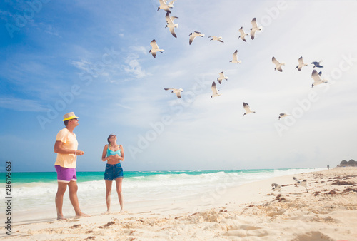Couple feeding birds on the sea shore, gulls flying over two persons walking by the sea