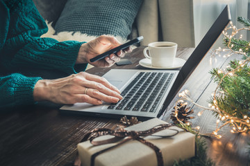 Christmas sales. Woman shopping with smartphone by laptop in home interior. Xmas. Planning holidays.