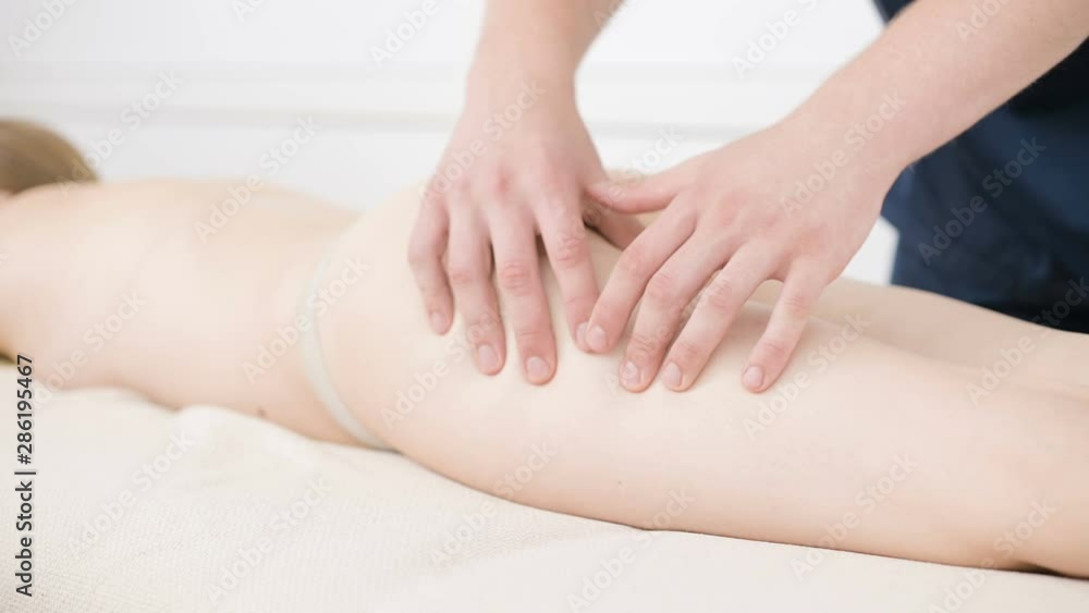 Close-up of a male physiotherapist doing a light massage to warm up the hips and buttocks of a young woman in a professional spa salon next to a sprig of cotton. 4K massage body care concept Close-up of a male physiotherapist doing a light massage to warm up the hips and buttocks of a young woman in a professional spa salon next to a sprig of cotton. 4K massage body care concept