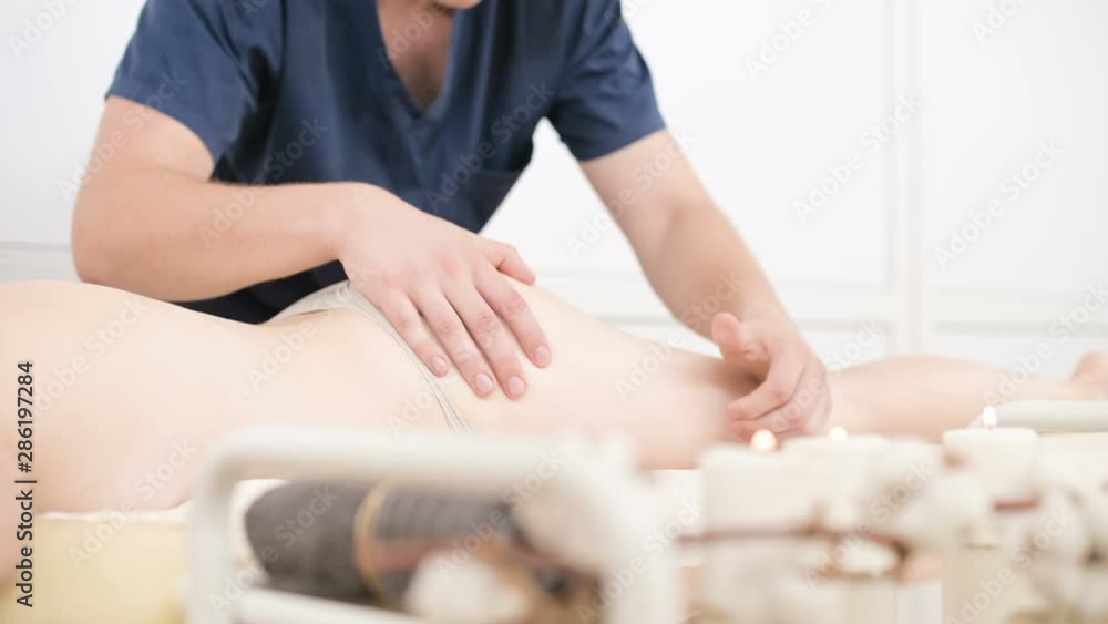 Close-up of a male physiotherapist doing a light massage to warm up the hips and buttocks of a young woman in a professional spa salon next to a sprig of cotton. 4K massage body care concept Close-up of a male physiotherapist doing a light massage to warm up the hips and buttocks of a young woman in a professional spa salon next to a sprig of cotton. 4K massage body care concept