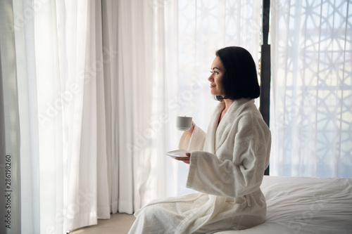 Woman wearing white bathrobe standing at morning near window in the light room