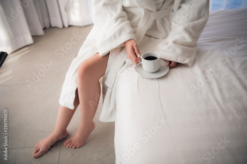 Close-up of legs of woman in white terry bathrobe on bed with cup of coffee in the morning. Ideal figure in light room. photo