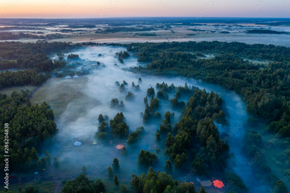 Naklejka premium Aerial view of the forest, field and river covered with layers of thick morning fog